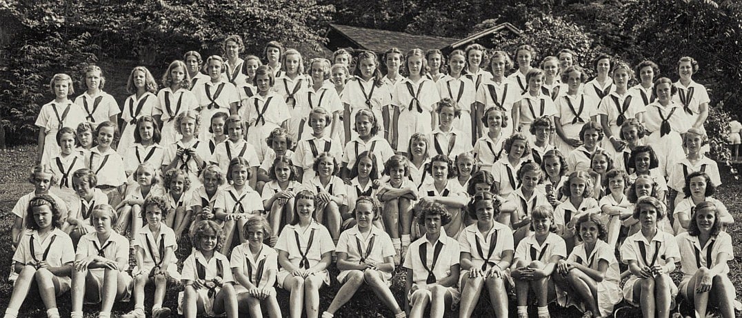 A large group of girls in matching uniforms with neckerchiefs poses outdoors in several rows on grassy steps with trees in the background.