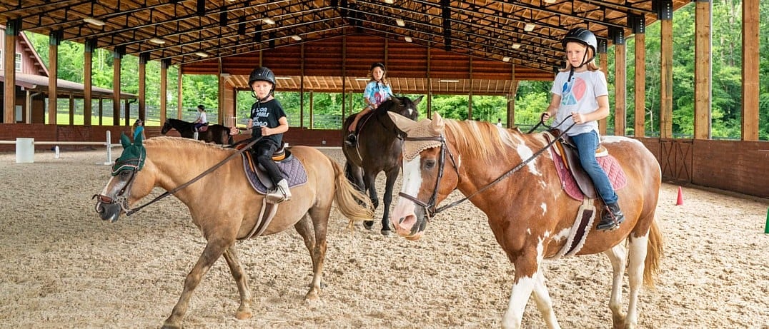 Three children wearing helmets ride horses at a covered riding arena during a lesson.