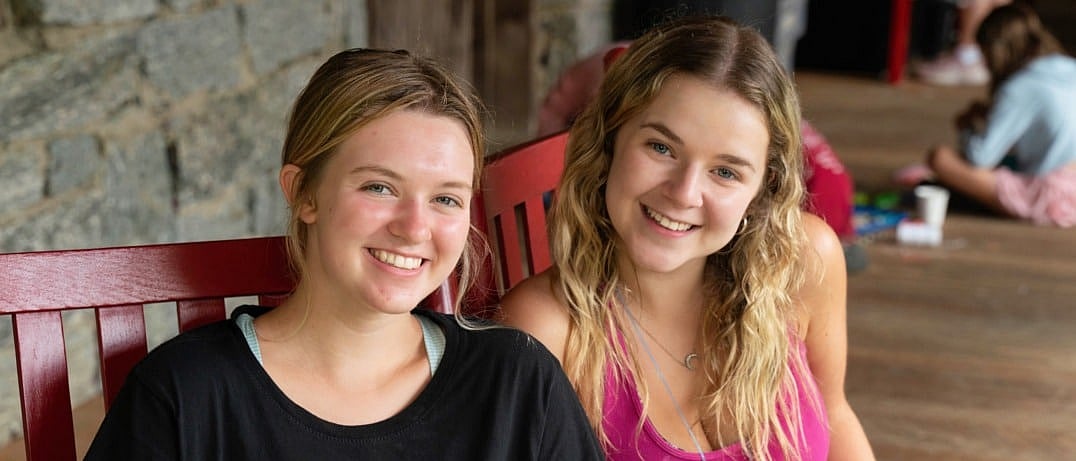 Two smiling girls sitting on a red wooden bench against a stone wall.