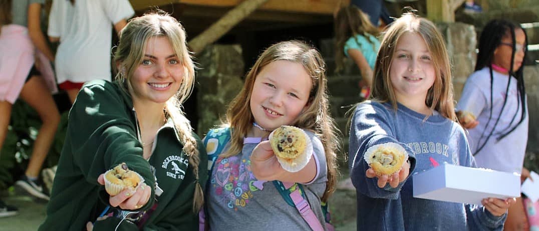 Three smiling girls holding out muffins towards the camera, with other children and outdoor steps in the background.