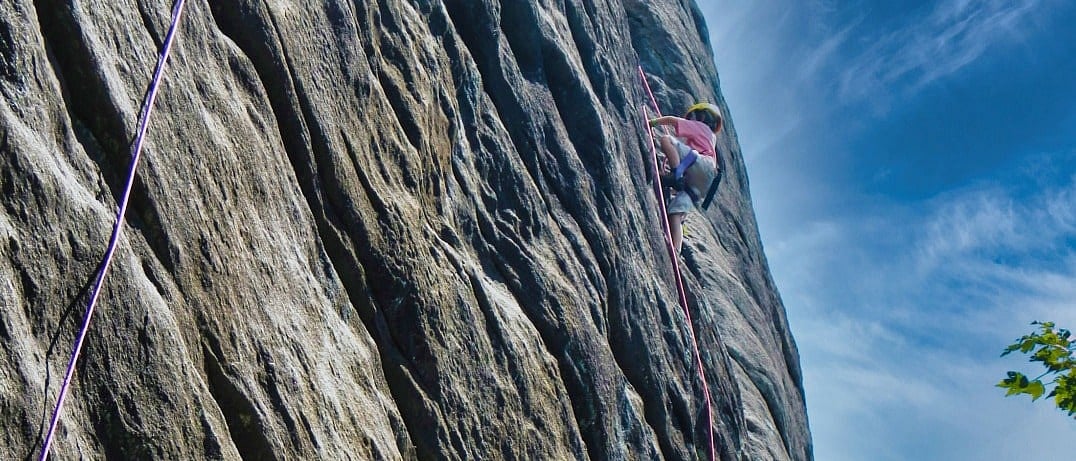 A child wearing a helmet climbs a steep rock face using a red safety rope under a blue sky.