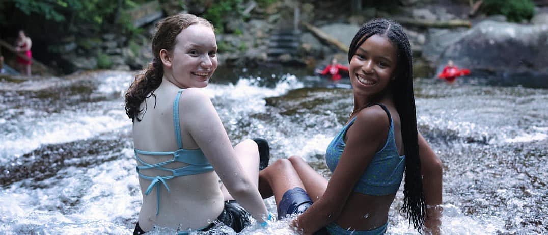 Two teenagers in swimwear smile at the camera while sitting and sliding in a shallow, flowing river.