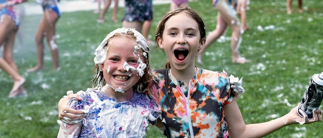 Two campers smiling and covered in shaving cream while enjoying an outdoor group activity on the grass.