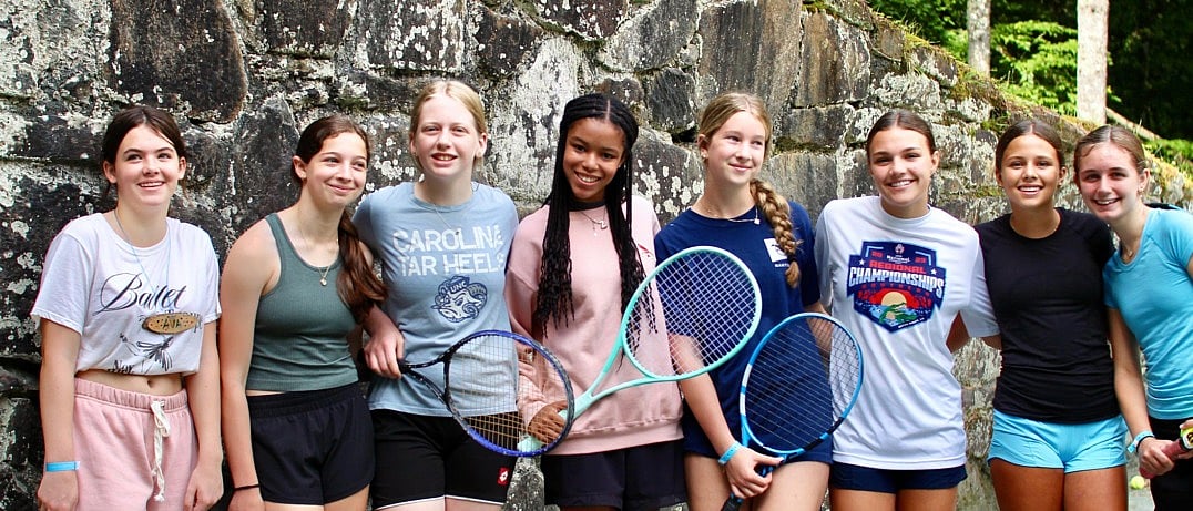 A group of eight teens stand closely together outdoors, holding tennis rackets and smiling in front of a stone wall.