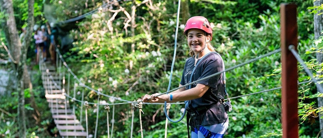 A child wearing a red helmet and safety harness smiles while crossing a rope bridge surrounded by green forest.