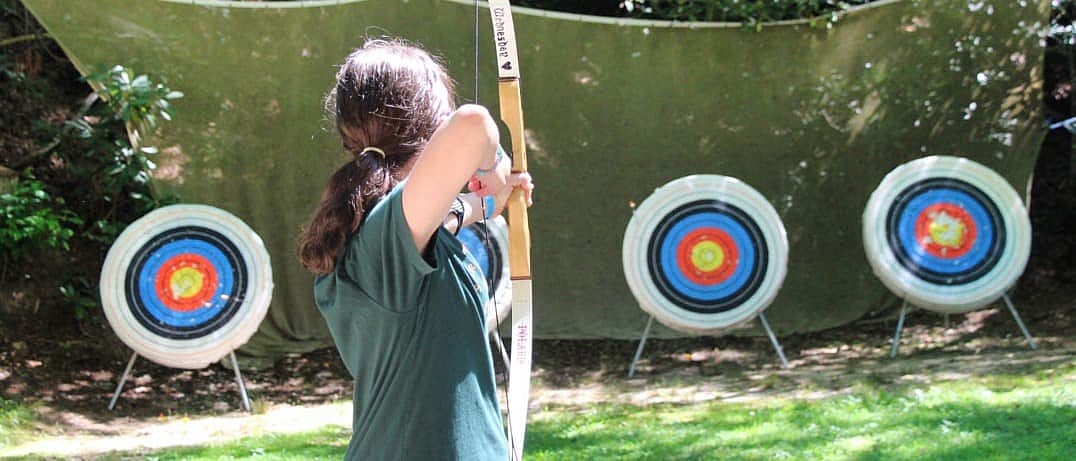 A camp girl is aiming a wooden bow at three archery targets set up outdoors with a green backdrop.