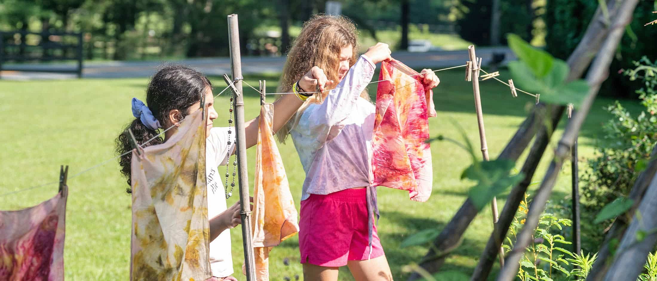 Two campers hang vibrant, naturally dyed fabrics on a clothesline outdoors on a sunny day.