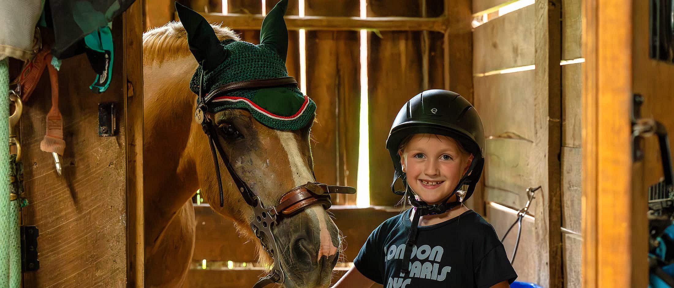 A camper wearing a helmet stands beside a horse in a wooden stable, smiling while gently holding the horse’s bridle.