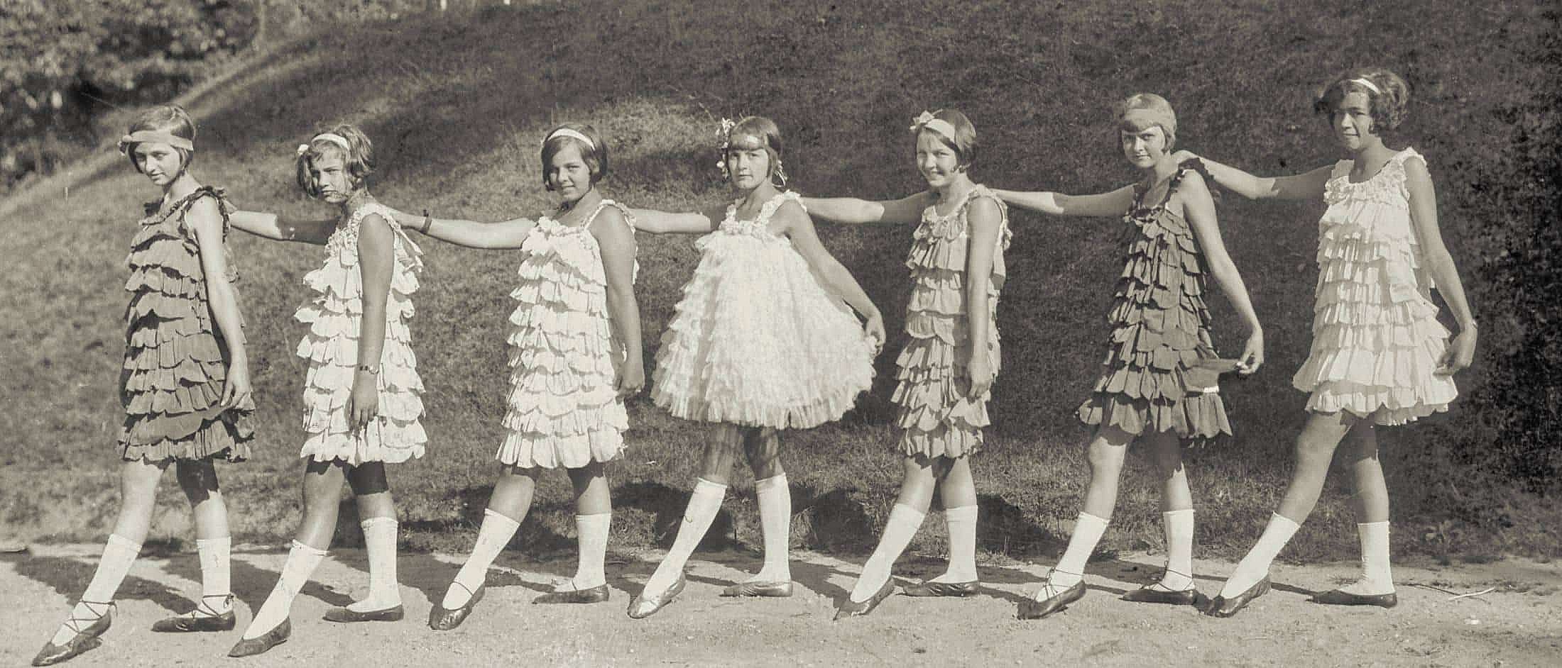 Seven girls wearing ruffled flapper-style dresses stand in a line outdoors with arms resting on each other's shoulders and one leg extended forward.