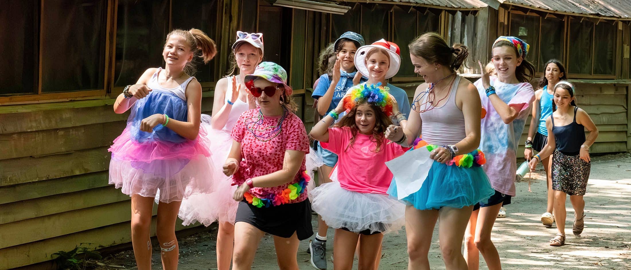 A group of children wearing colorful tutus, hats, and playful accessories walk together along a shaded wooden cabin path.