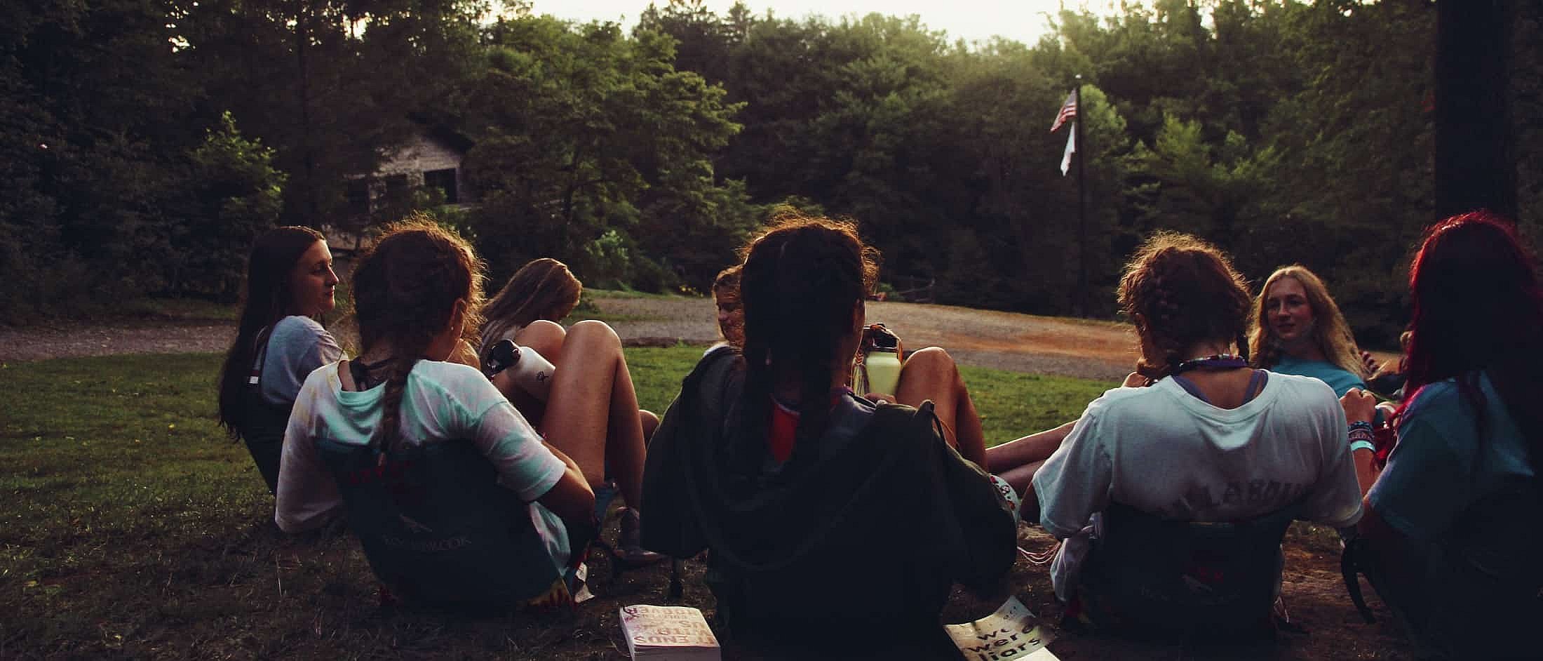 A group of girls sitting in a circle on the grass outdoors, surrounded by trees and conversing during early evening.