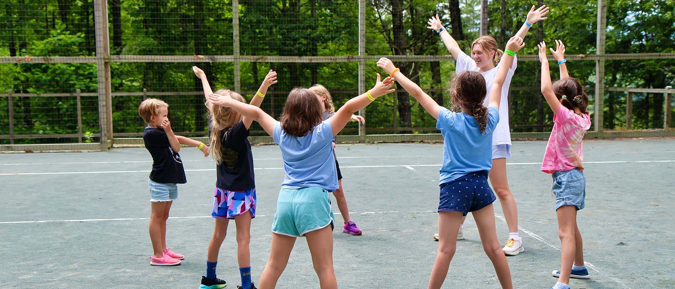 A group of children standing in a circle on a tennis court with their arms raised, led by an instructor.