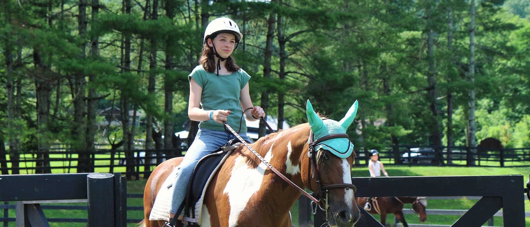 A camper wearing a helmet rides a brown and white horse with a teal ear bonnet for a lesson in an outdoor riding area surrounded by trees.