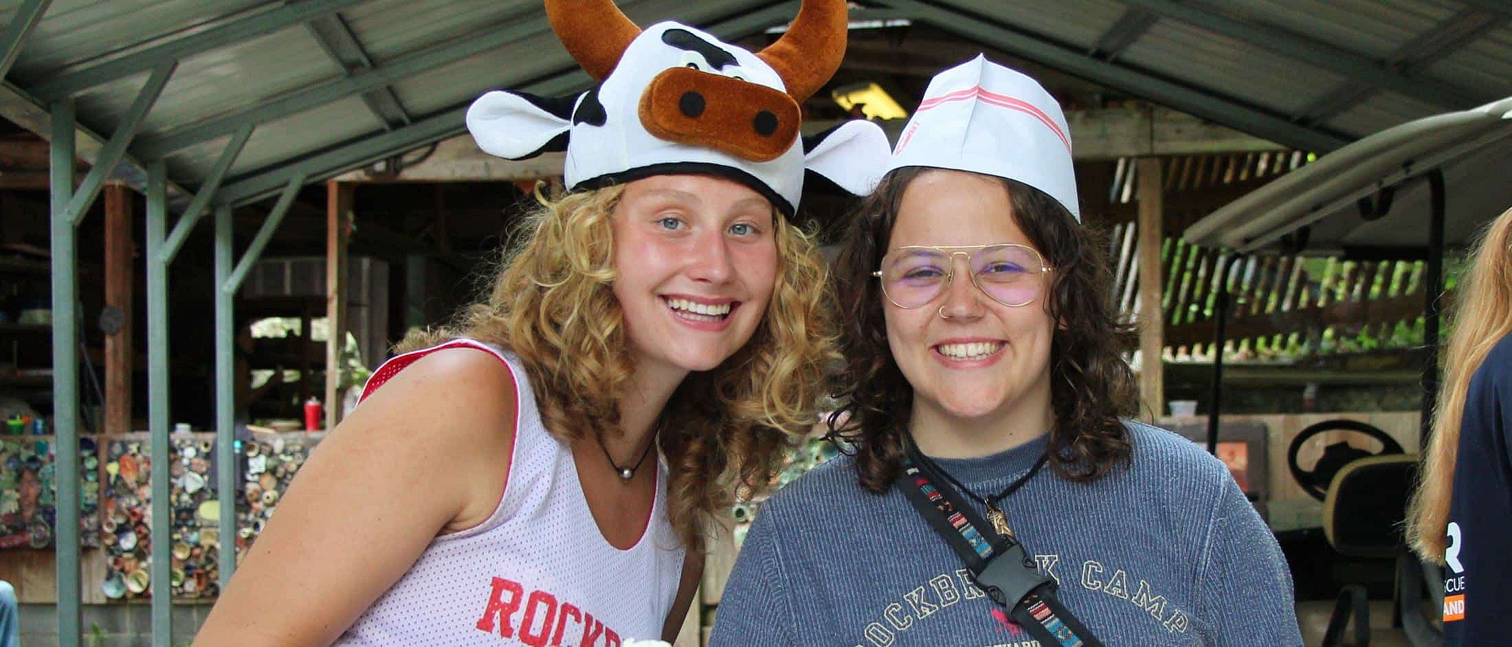 Two smiling counselors pose together, one wearing a cow-themed hat and the other wearing a white paper hat under a covered outdoor area.