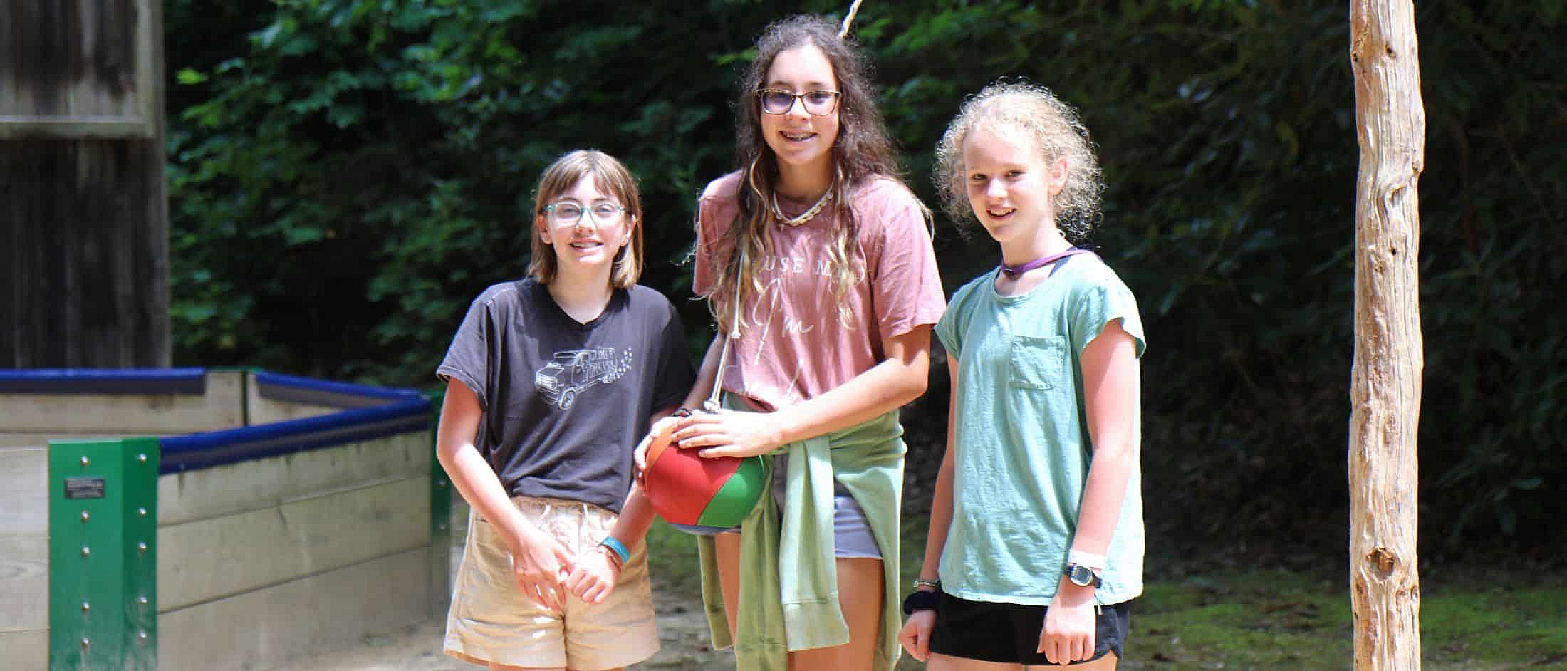 Three girls stand together near a tetherball pole, with one girl holding the tetherball.