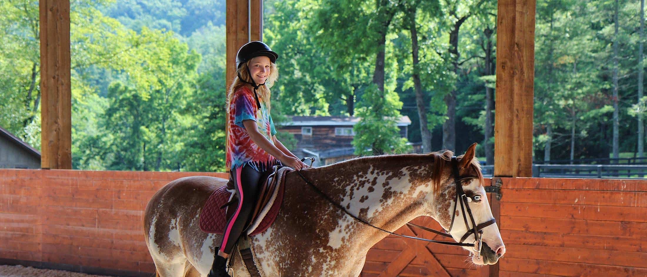 Teen rider wearing a helmet sits on a speckled horse inside a wooden riding arena with trees visible outside.