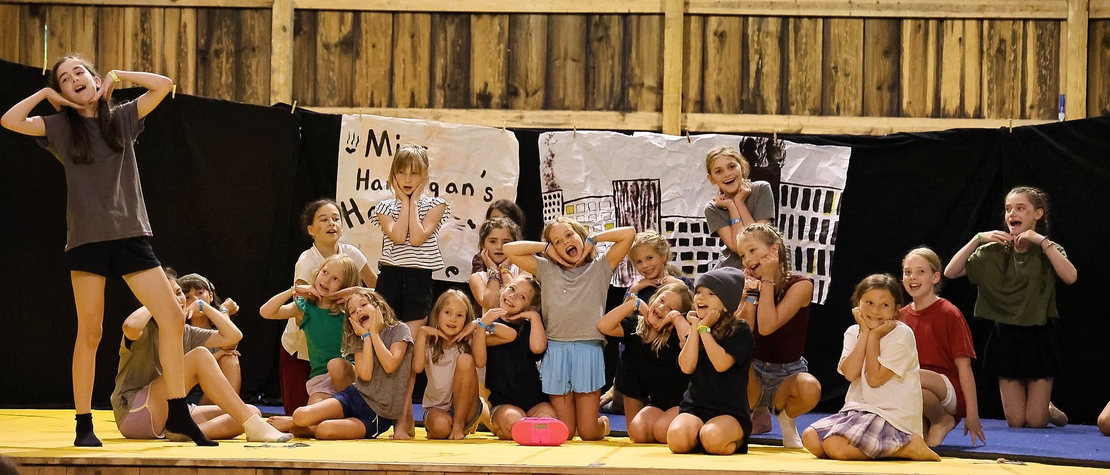 A group of campers on stage performing a musical with expressive faces and hand gestures in front of hand-drawn backdrop signs.