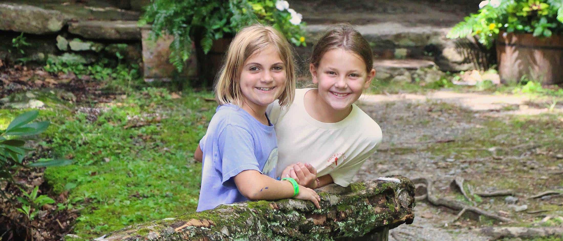 Two girls smiling and leaning on a moss-covered wooden fence in a forested area.