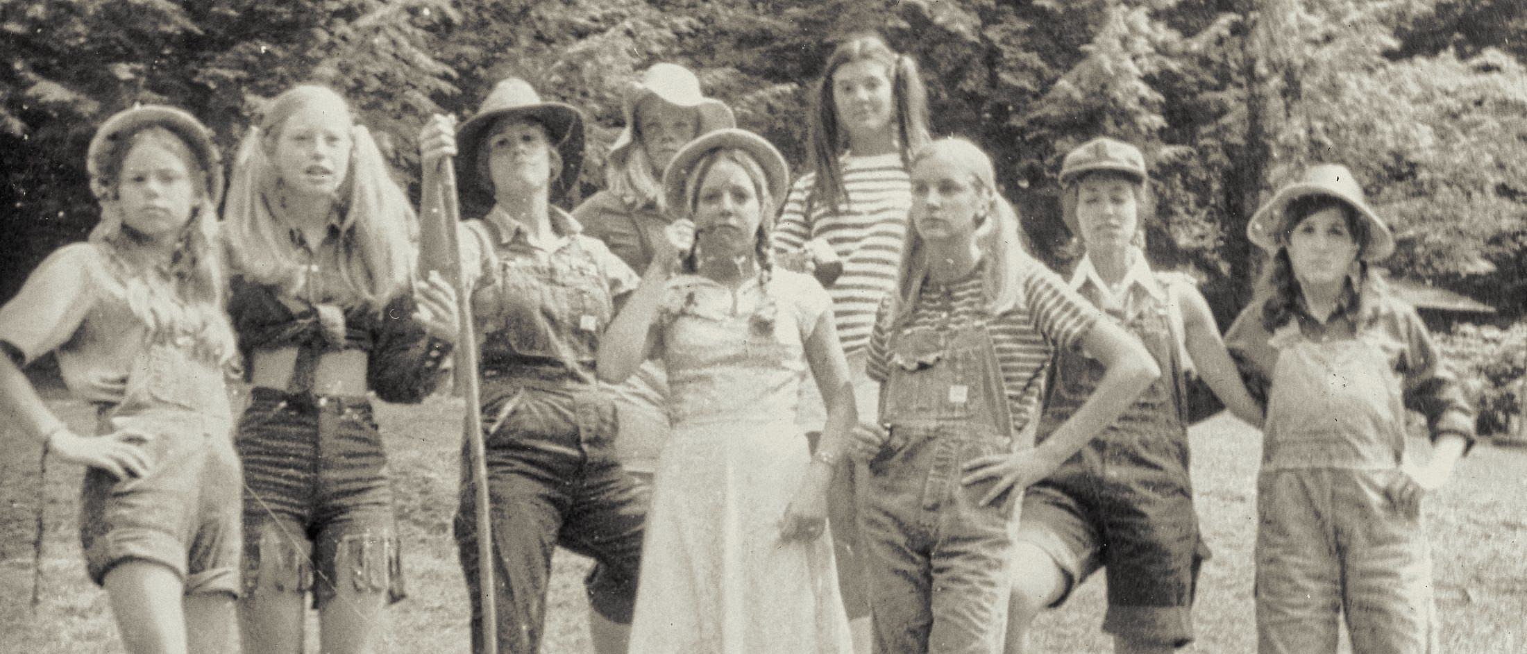 A group of campers in vintage 1940s-style clothing and hats pose outdoors with confident stances against a leafy background.