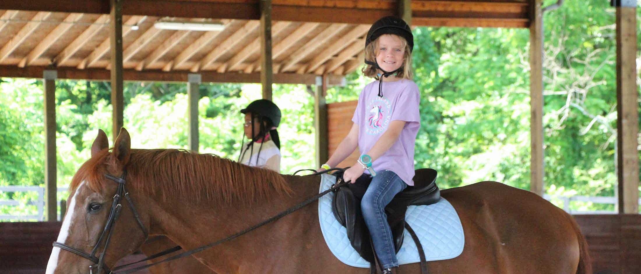 A smiling camper wearing a helmet sits on a brown horse inside a covered riding arena with another rider nearby.