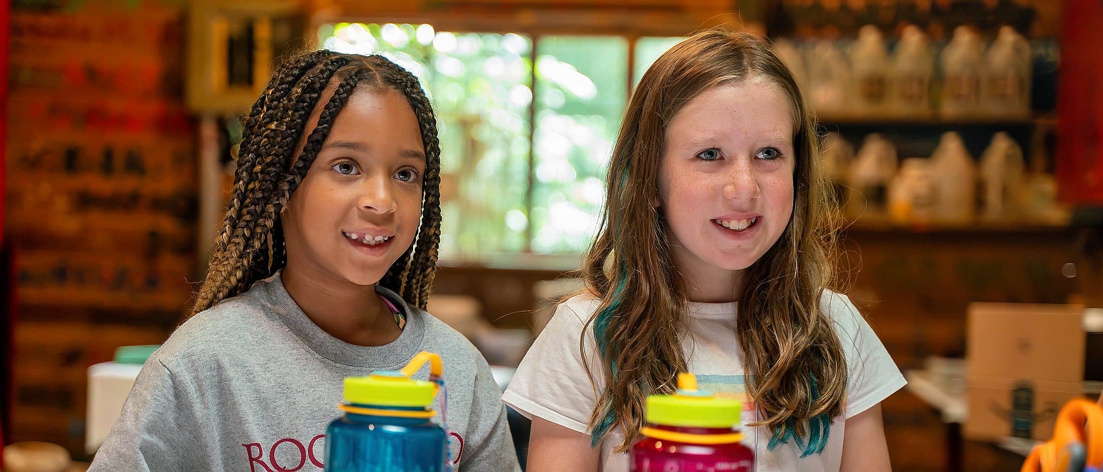 Two girls sit indoors at a table with colorful water bottles, appearing engaged in a craft or camp activity.