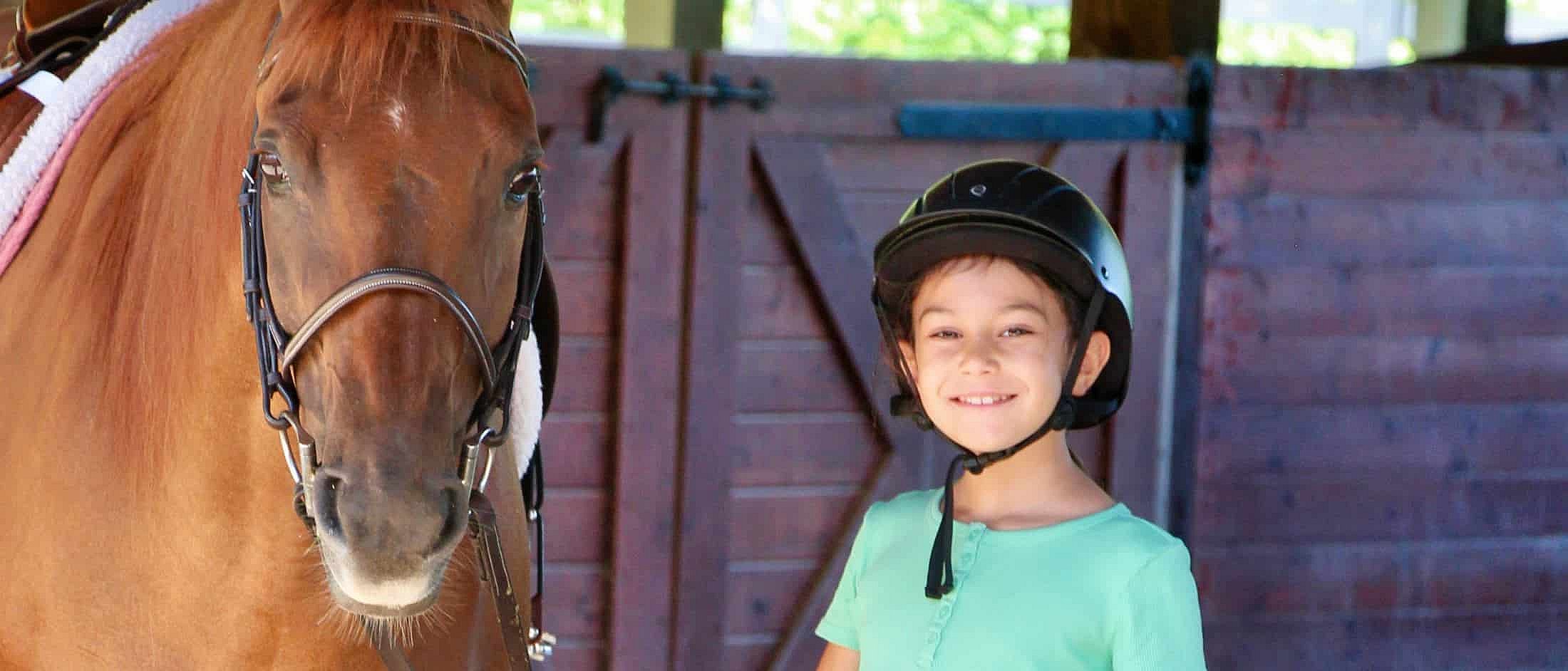 A smiling child wearing a riding helmet stands next to a brown horse inside a wooden stable.