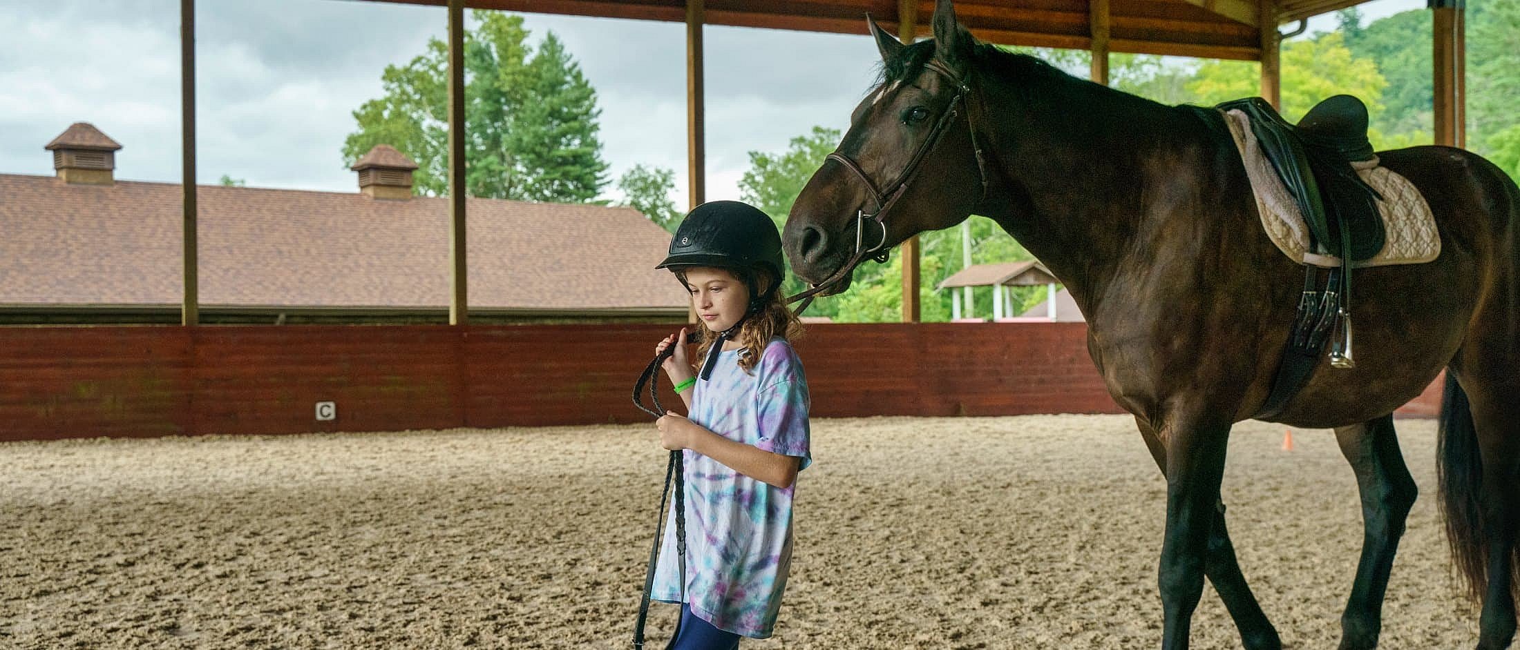 A girl wearing a helmet leads a saddled horse by the reins inside a covered riding arena.