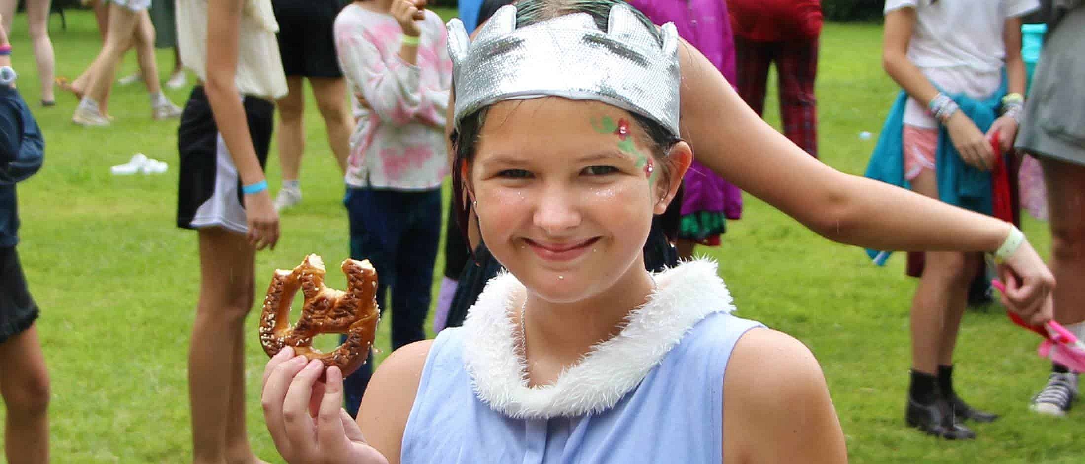 A smiling child wearing a silver crown and light costume holds a partially eaten pretzel during an outdoor gathering on grass.