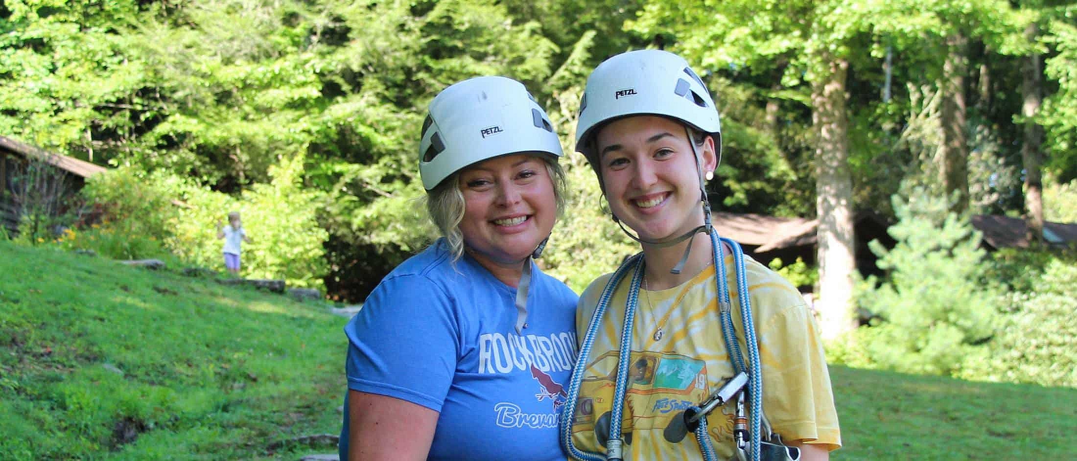 Two people wearing white climbing helmets smile together outdoors with trees and rustic buildings in the background.