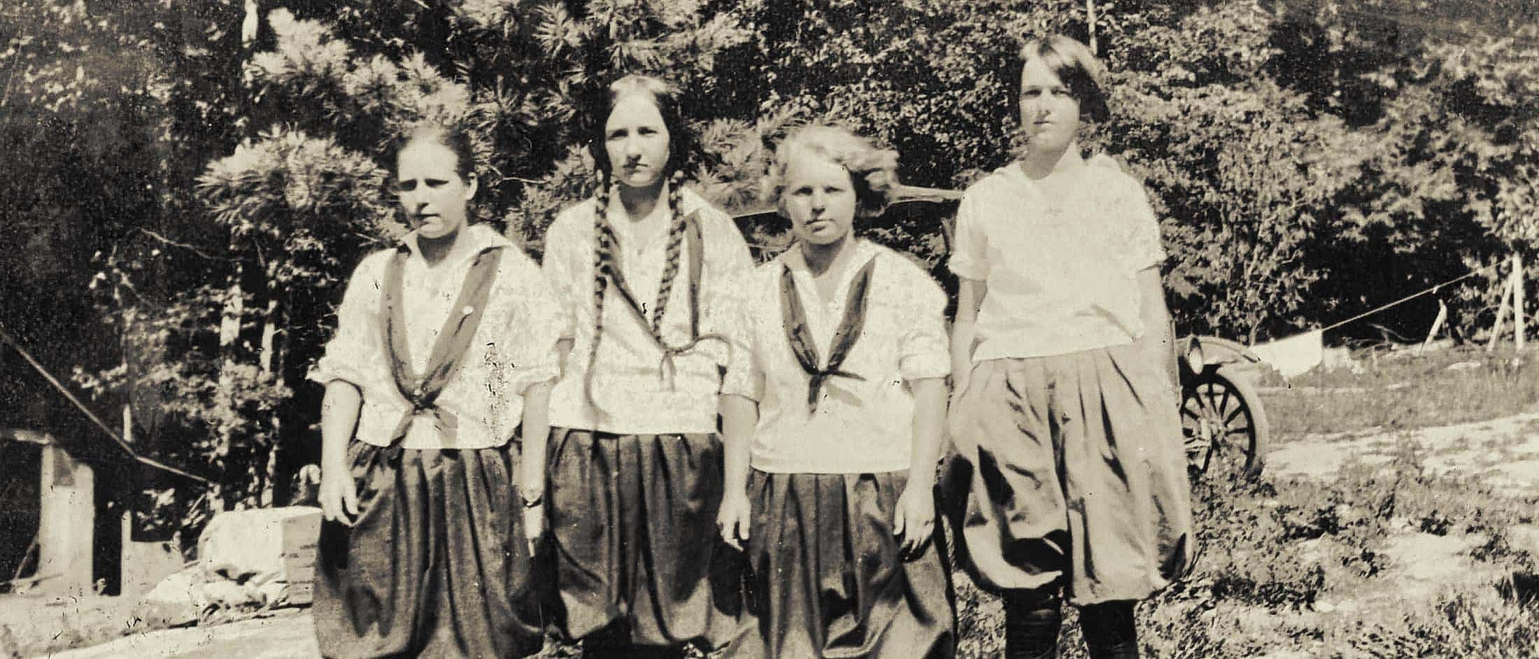 Four girls wearing early 20th-century camp uniforms with blouses and long skirts stand outdoors in front of trees and a vintage car.