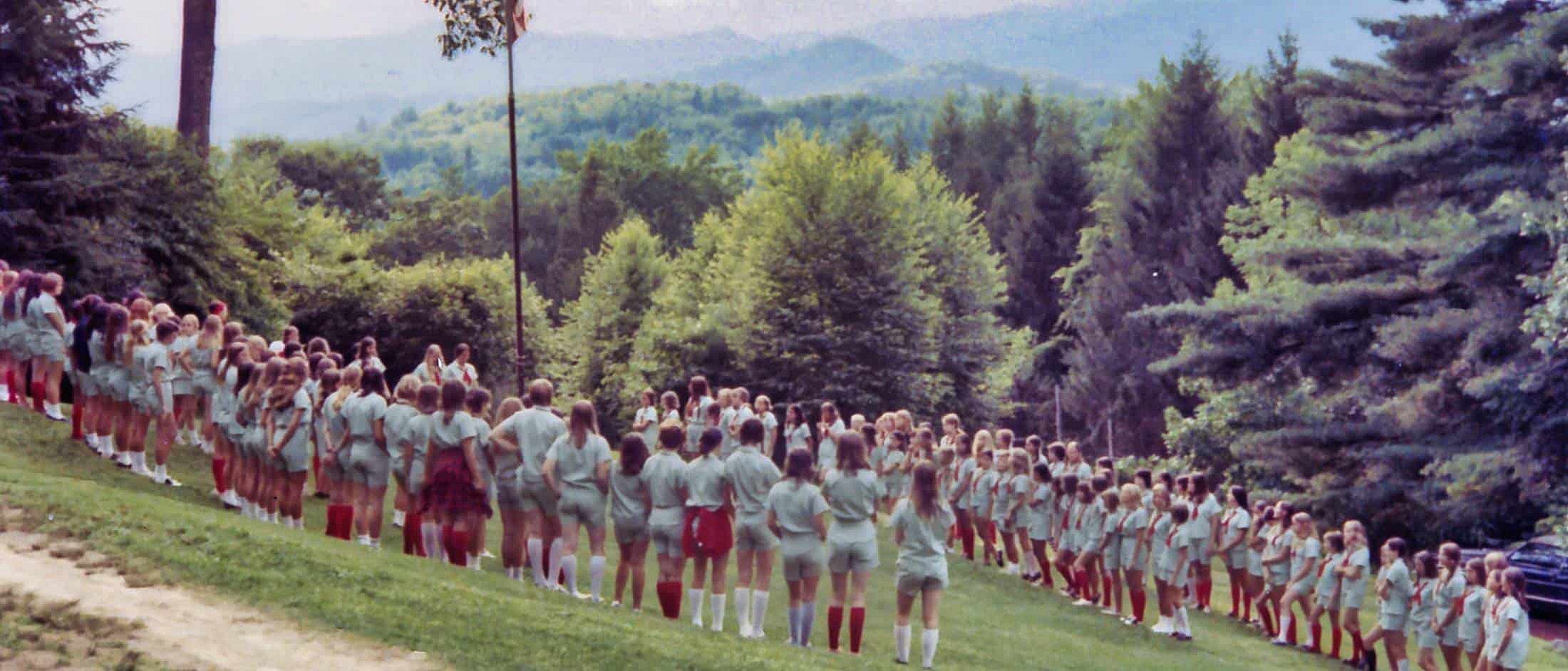 Large group of campers standing in rows on a grassy hillside surrounded by trees during an outdoor assembly.
