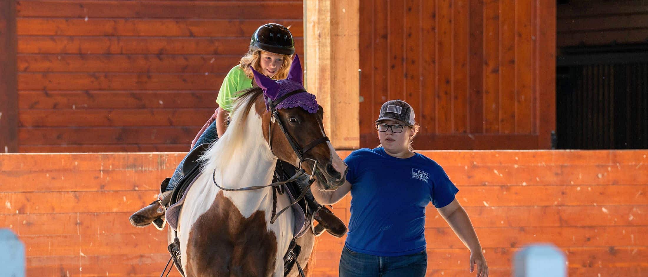 A child wearing a helmet rides a horse indoors while another person in a blue shirt leads the horse by its reins.