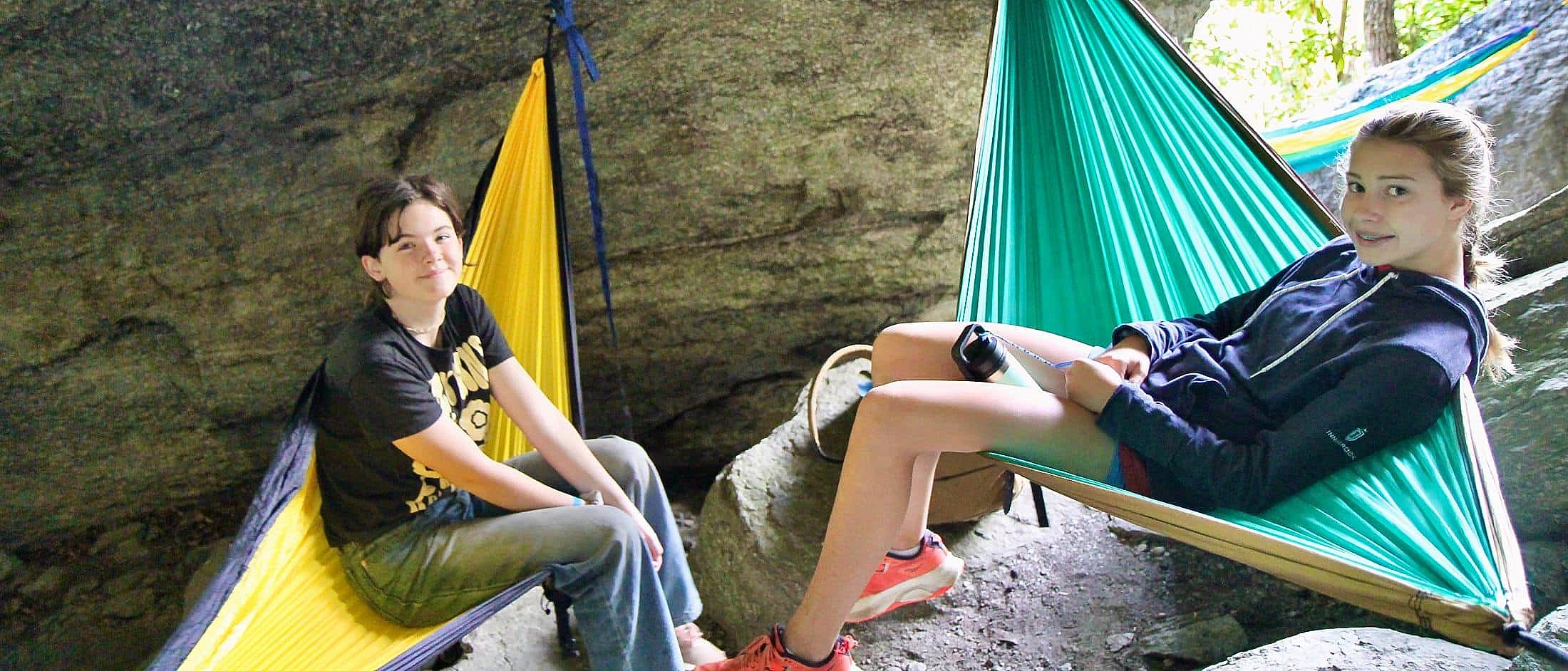 Two campers relax in colorful hammocks suspended between rocks in a shaded outdoor area.