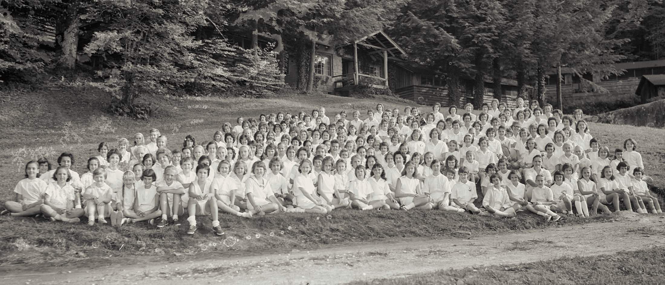 A large group of campers and counselors sit and stand on a grassy hillside in front of rustic wooden cabins surrounded by trees.