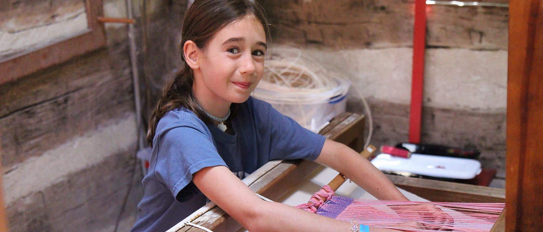 A young camper seated indoors weaving pink and purple threads on a wooden loom.