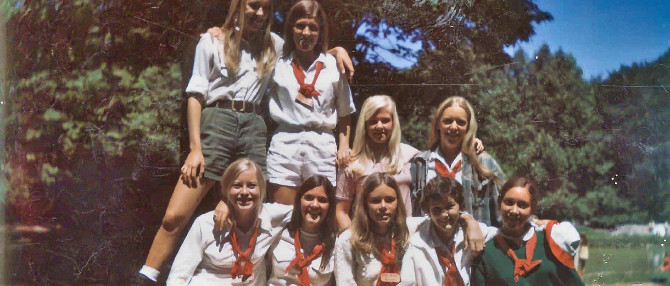 A group of nine campers in vintage summer clothing pose together outdoors in front of trees on a sunny day.