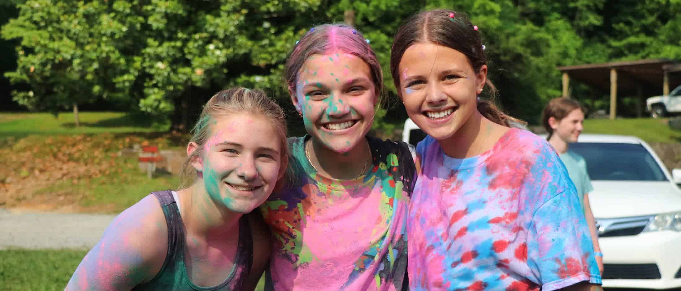 Three smiling campers covered in colorful powder pose outdoors on a sunny day with trees and cars in the background.