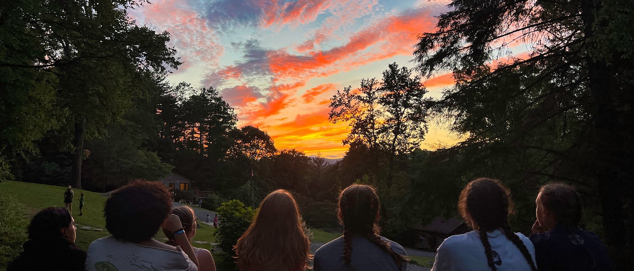 Campers sit on a hill facing a vibrant sunset sky with orange and pink clouds framed by tall trees.