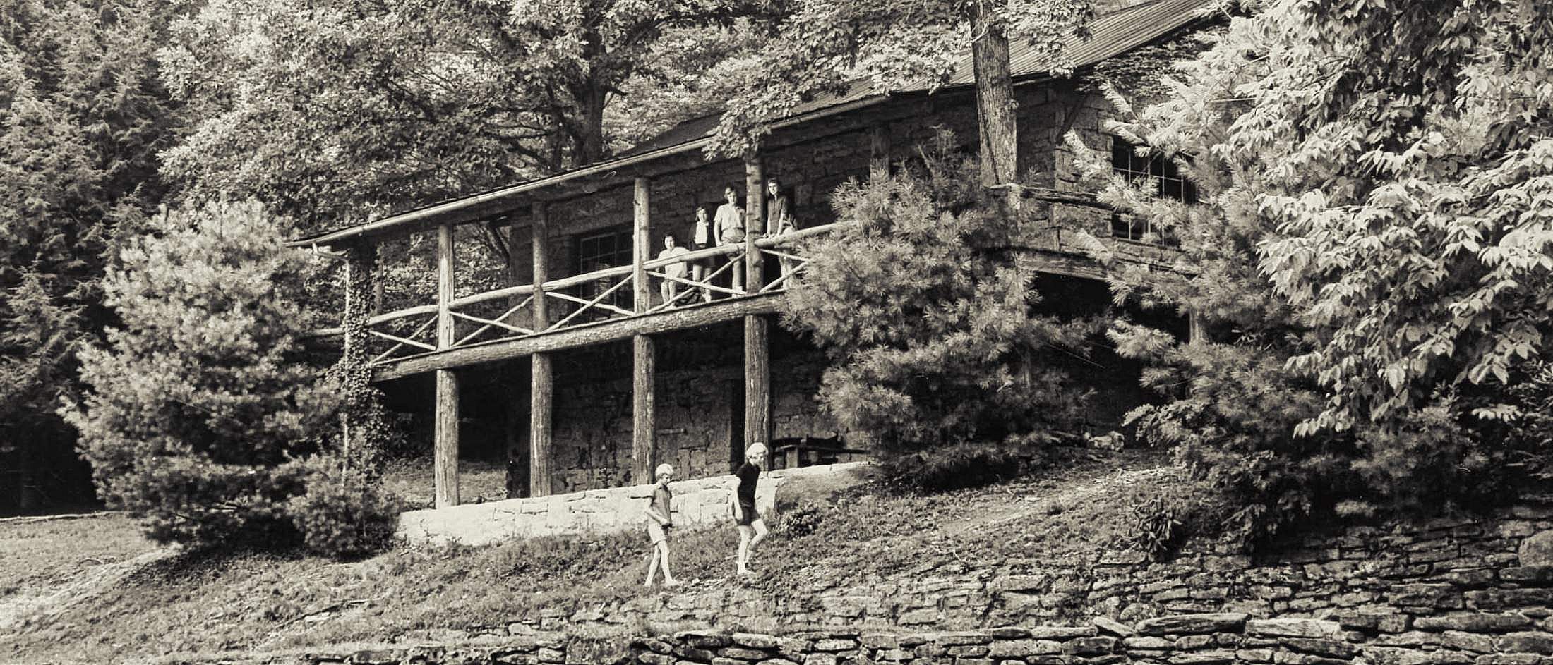 Two children walk along a stone wall in front of a rustic summer camp lodge surrounded by trees.