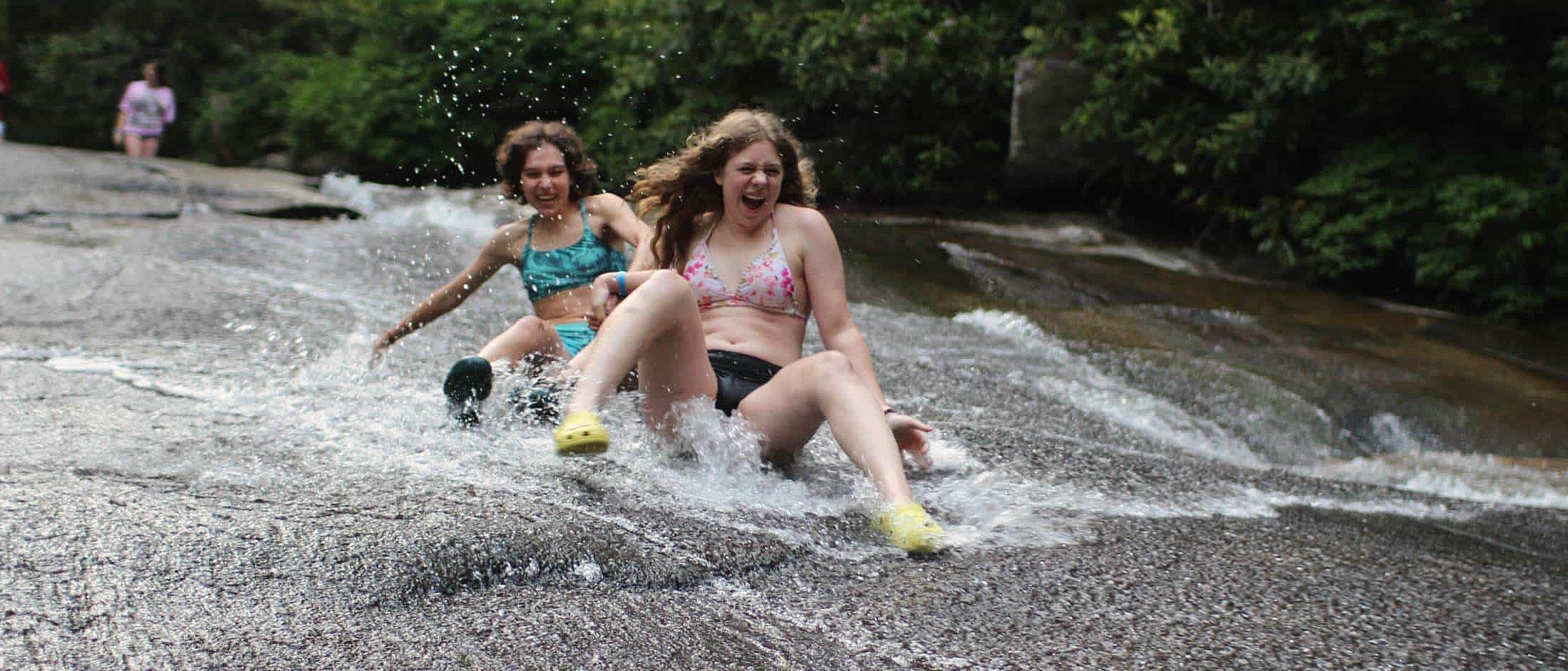 Two girls in swimsuits slide down a smooth rock surface with splashing water surrounded by forest.