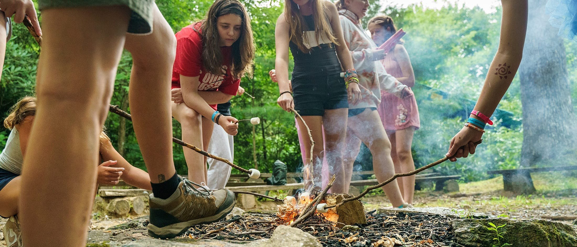 Campers gather around a small campfire outdoors, toasting marshmallows on sticks for making s'mores.