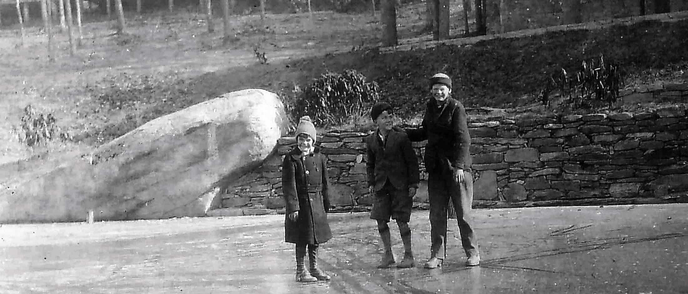 Three children stand on a frozen lake near a stone wall, dressed in warm coats, hats, and boots.
