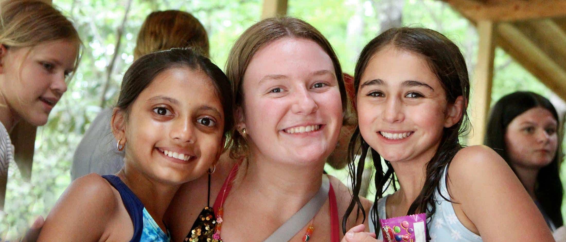 Three girls smiling closely together under a wooden shelter with other children visible in the background.