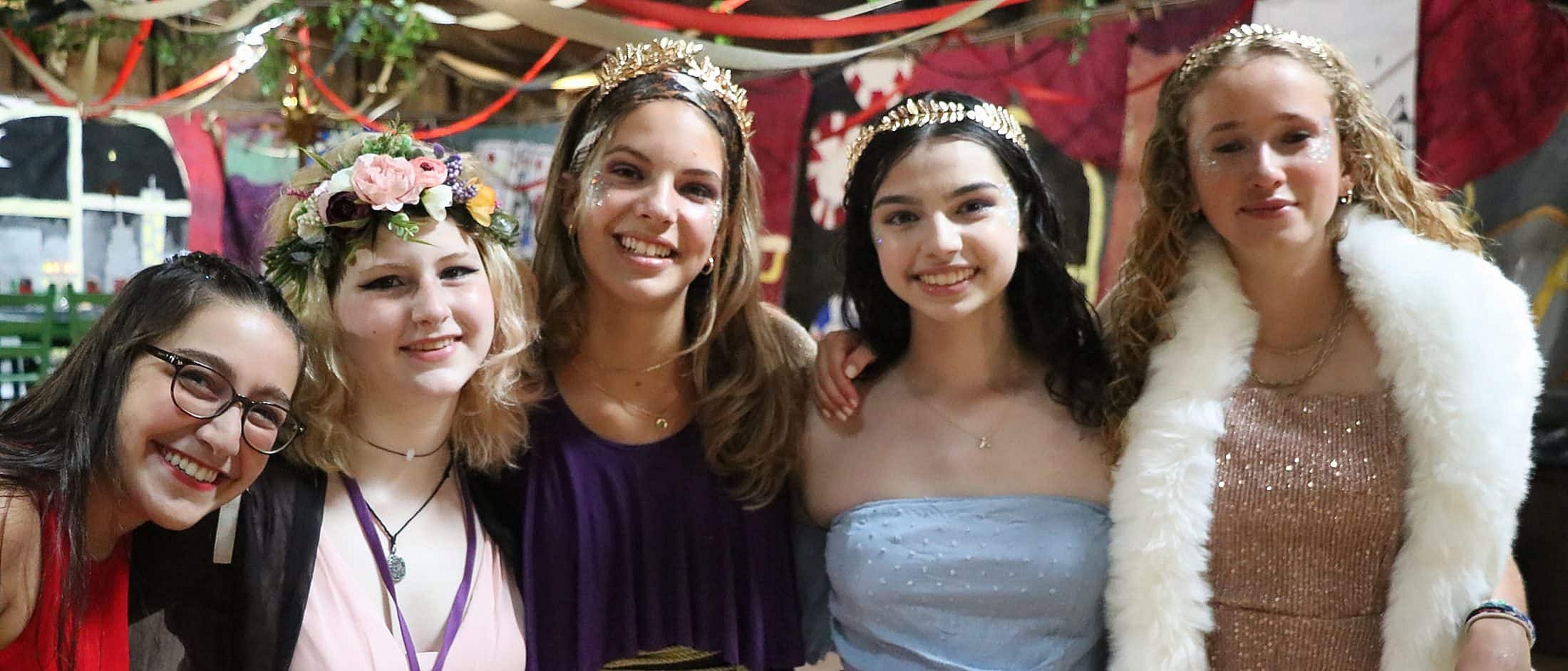 Five teens wearing festive costumes and headpieces stand closely together indoors, smiling with colorful decorations hanging in the background.