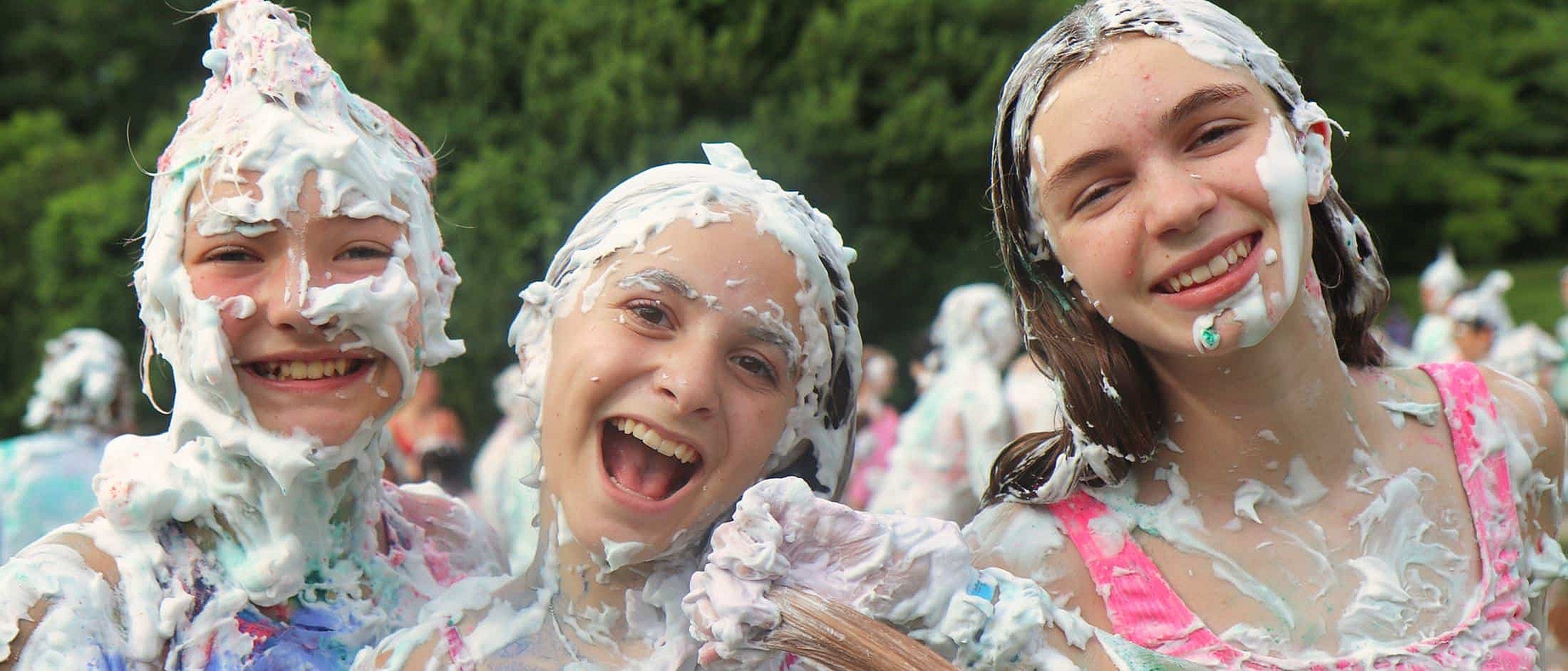Three teenagers smiling with their faces and hair covered in white foam outdoors with other people and trees in the background.