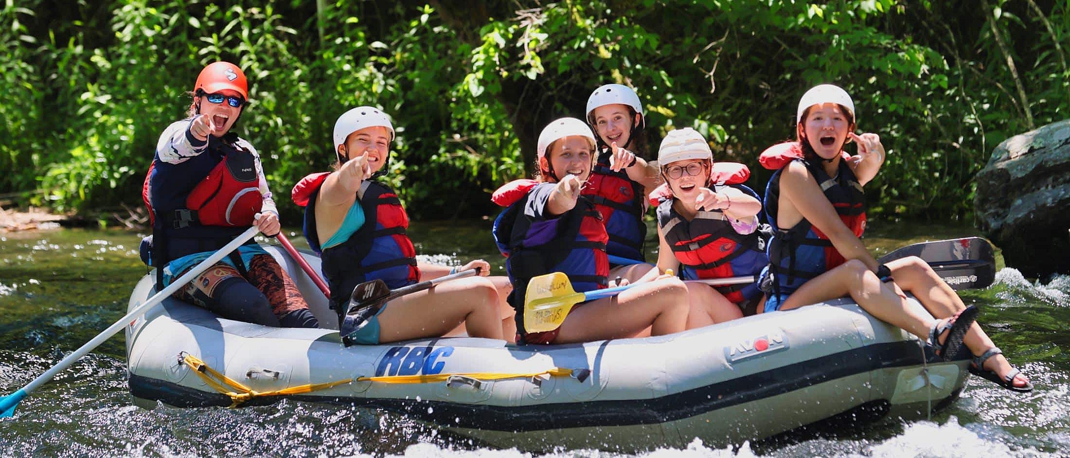 Six people wearing helmets and life jackets point and smile while sitting in a raft floating on a sunlit river with green foliage in the background.