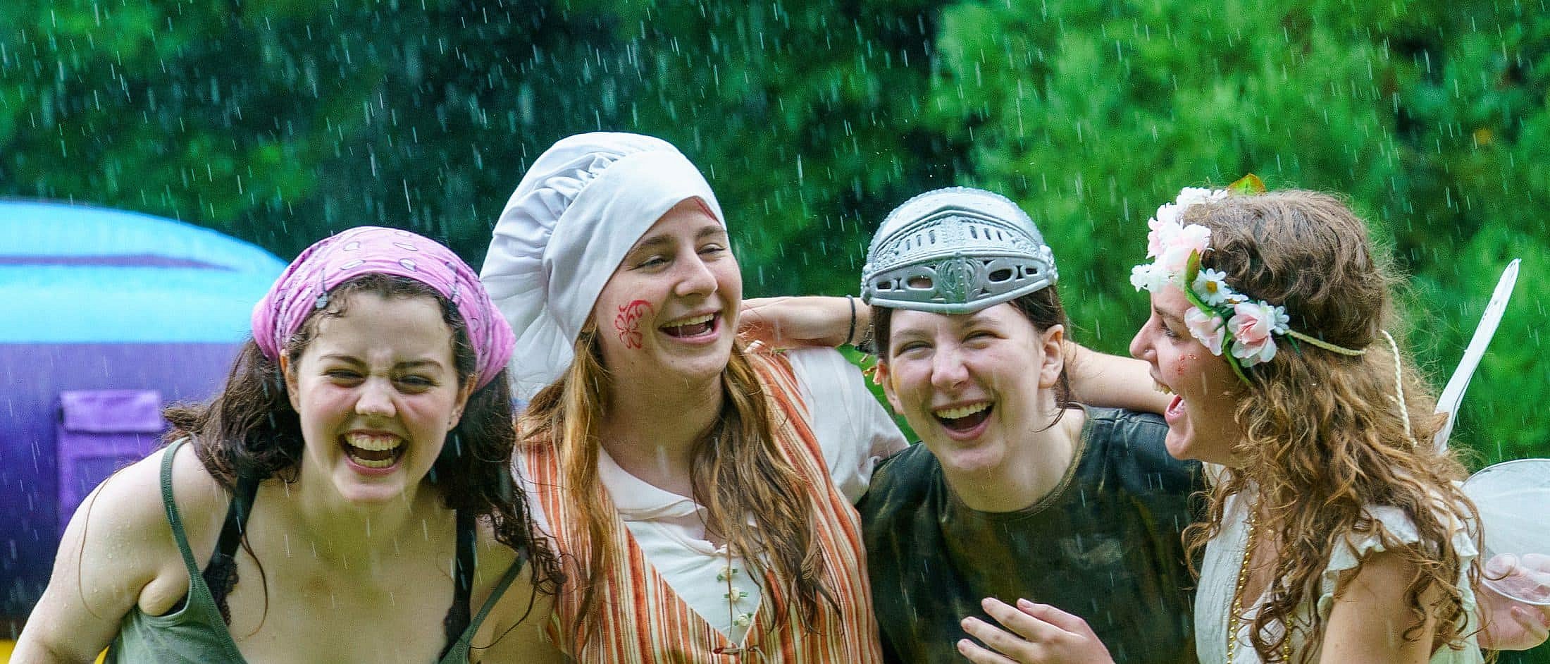 Four people wearing costumes and headpieces laugh together while standing outside in the rain.