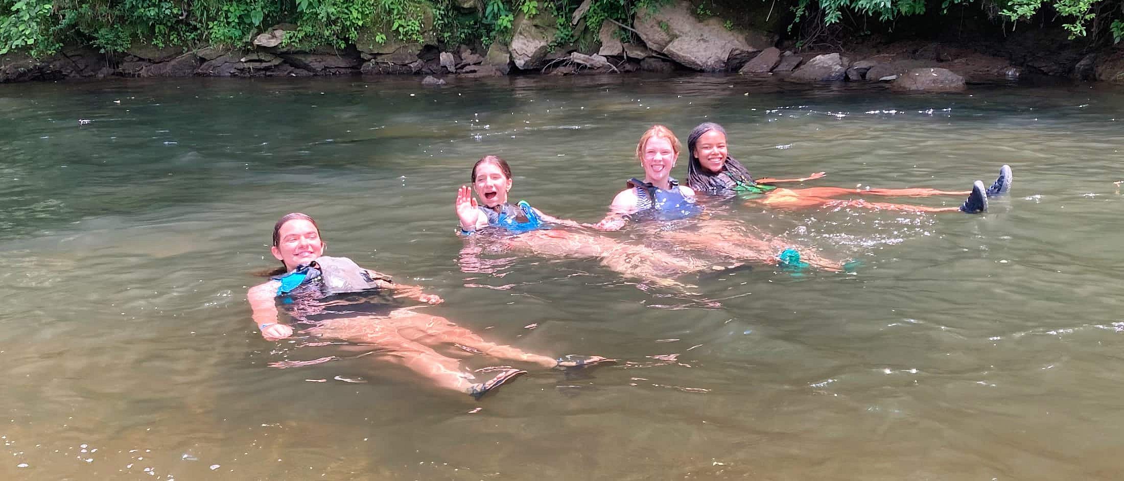 Four teens wearing life jackets are floating relaxed on their backs in a calm river surrounded by greenery.