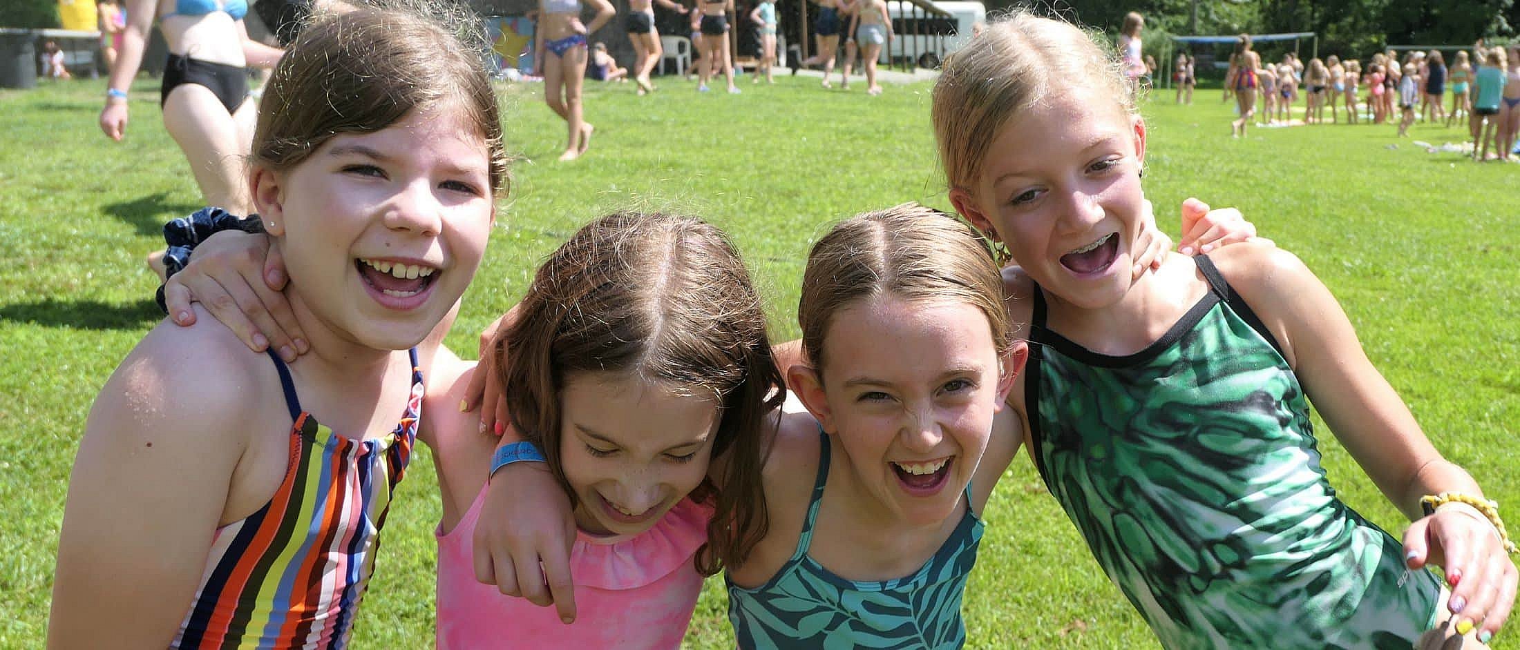 Four girls in colorful swimsuits stand closely together with arms around each other, smiling and laughing on a grassy field with other children playing in the background.