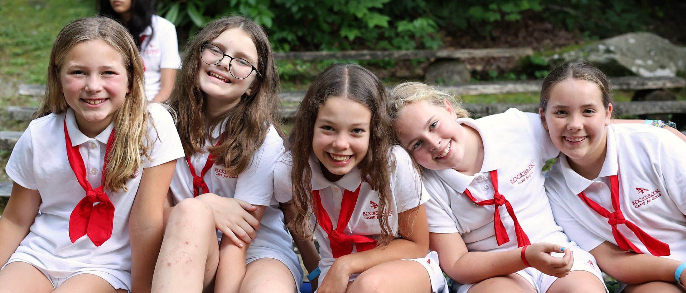 Five campers in white camp uniforms with red neckerchiefs sit closely together outdoors, smiling at the camera.